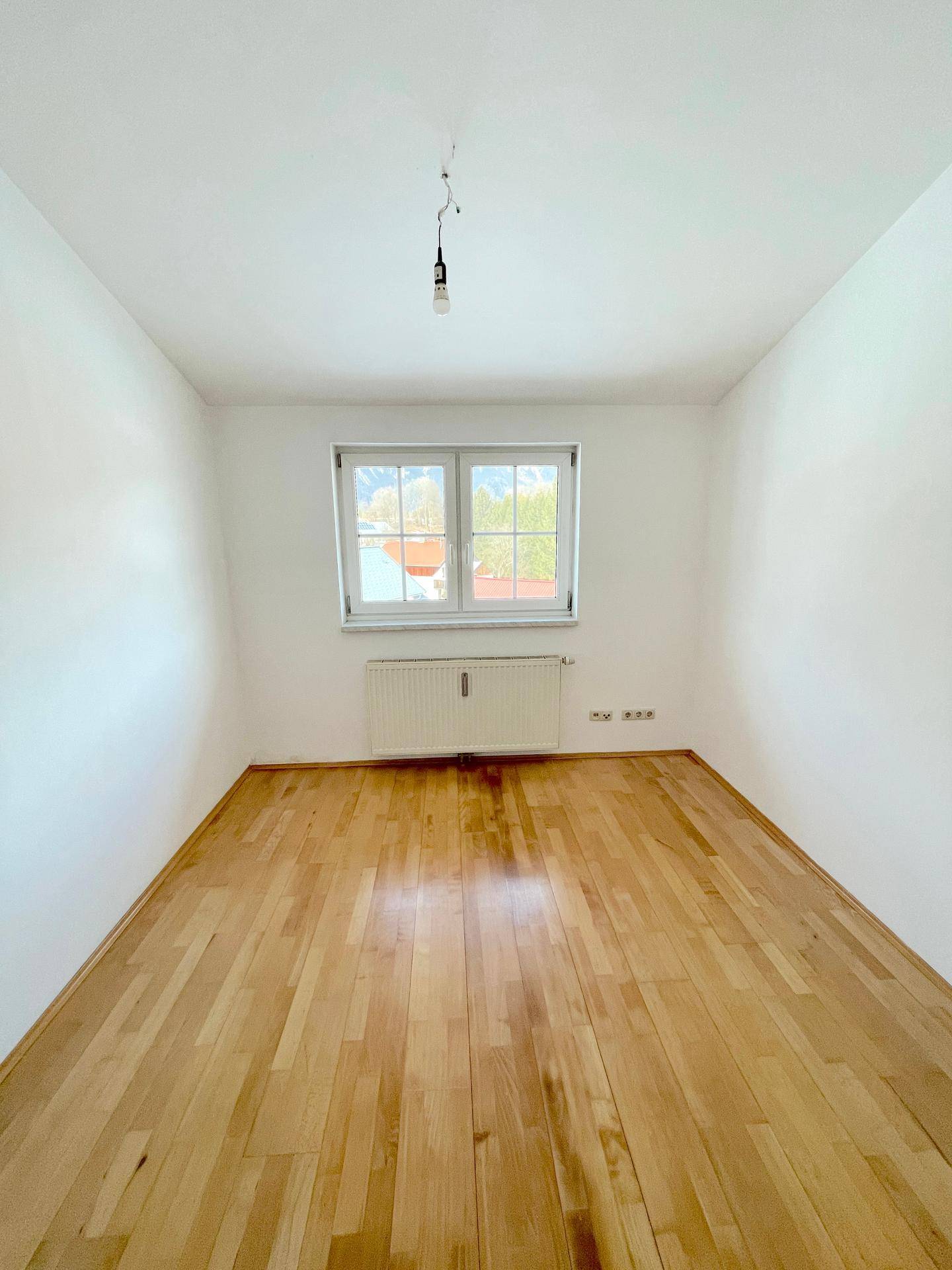 Helles und leeres Zimmer mit Holzboden und einem Fenster, das viel Tageslicht hereinlässt.