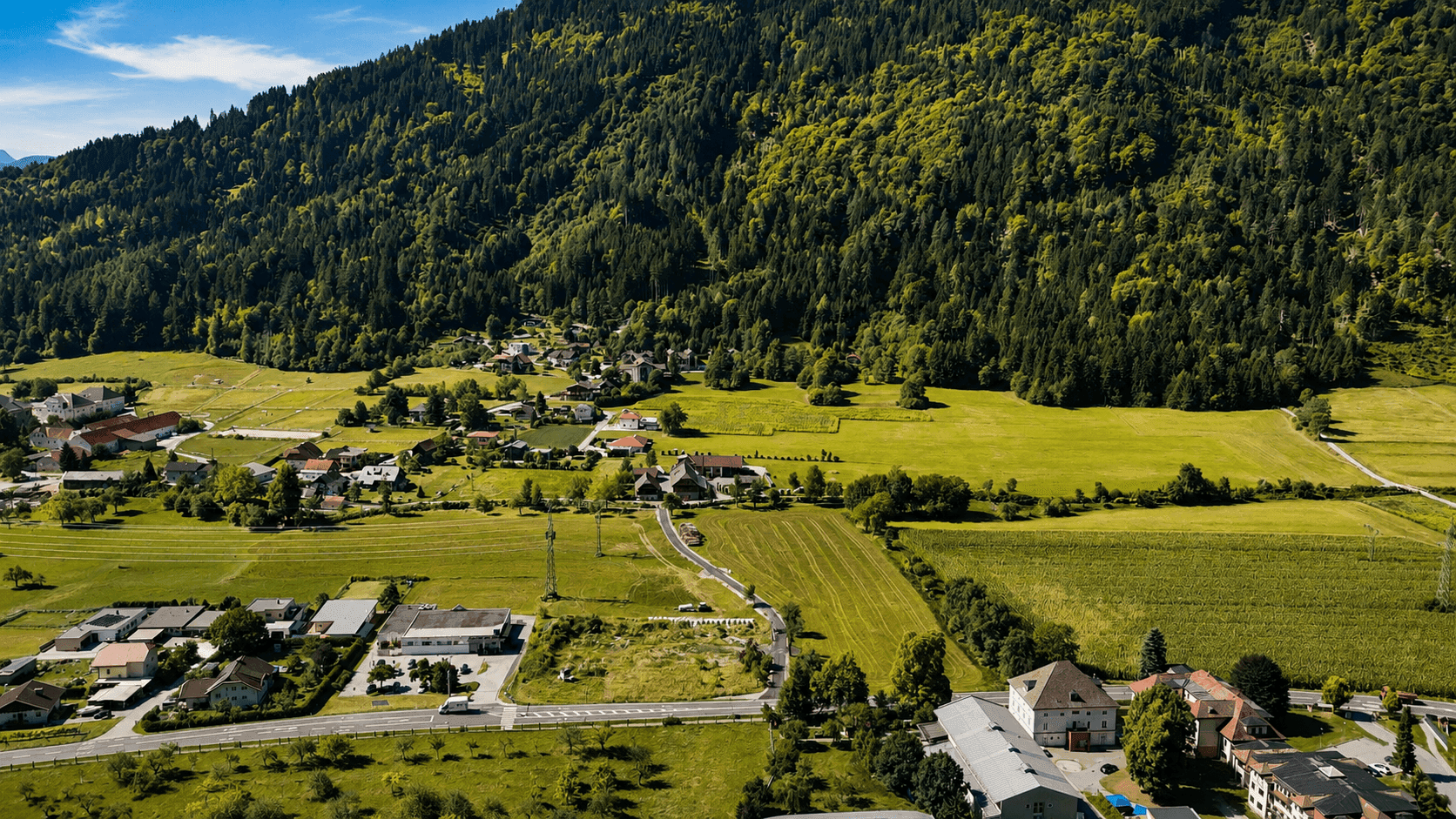 Weitläufige Luftaufnahme eines Tals mit bewaldeten Bergen, grünen Feldern und einer kleinen Siedlung.