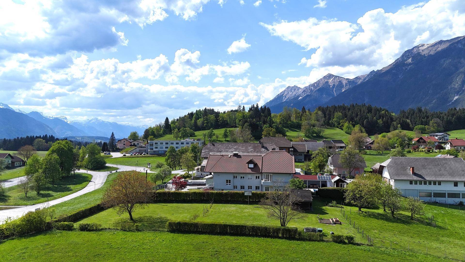 Panoramablick auf ein Wohnhaus in einer idyllischen Landschaft mit weiten Feldern und majestätischen Bergen unter blauem Himmel.