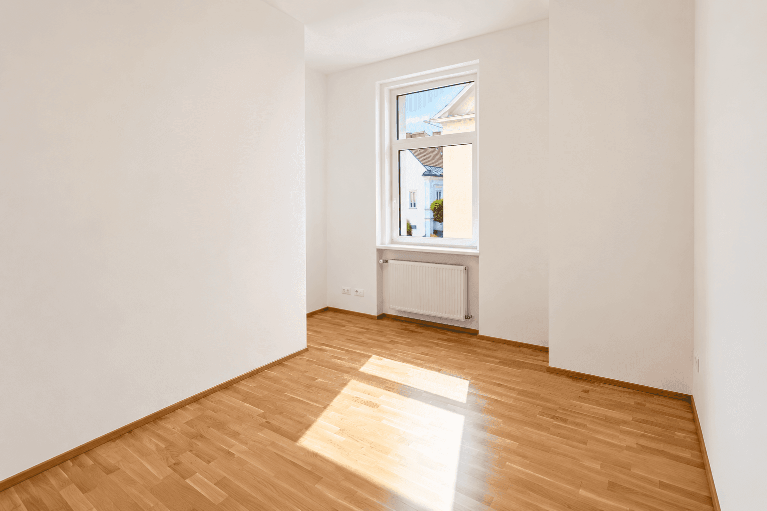 Freundliches Zimmer mit Parkettboden, einem Fenster und weiß gestrichenen Wänden.