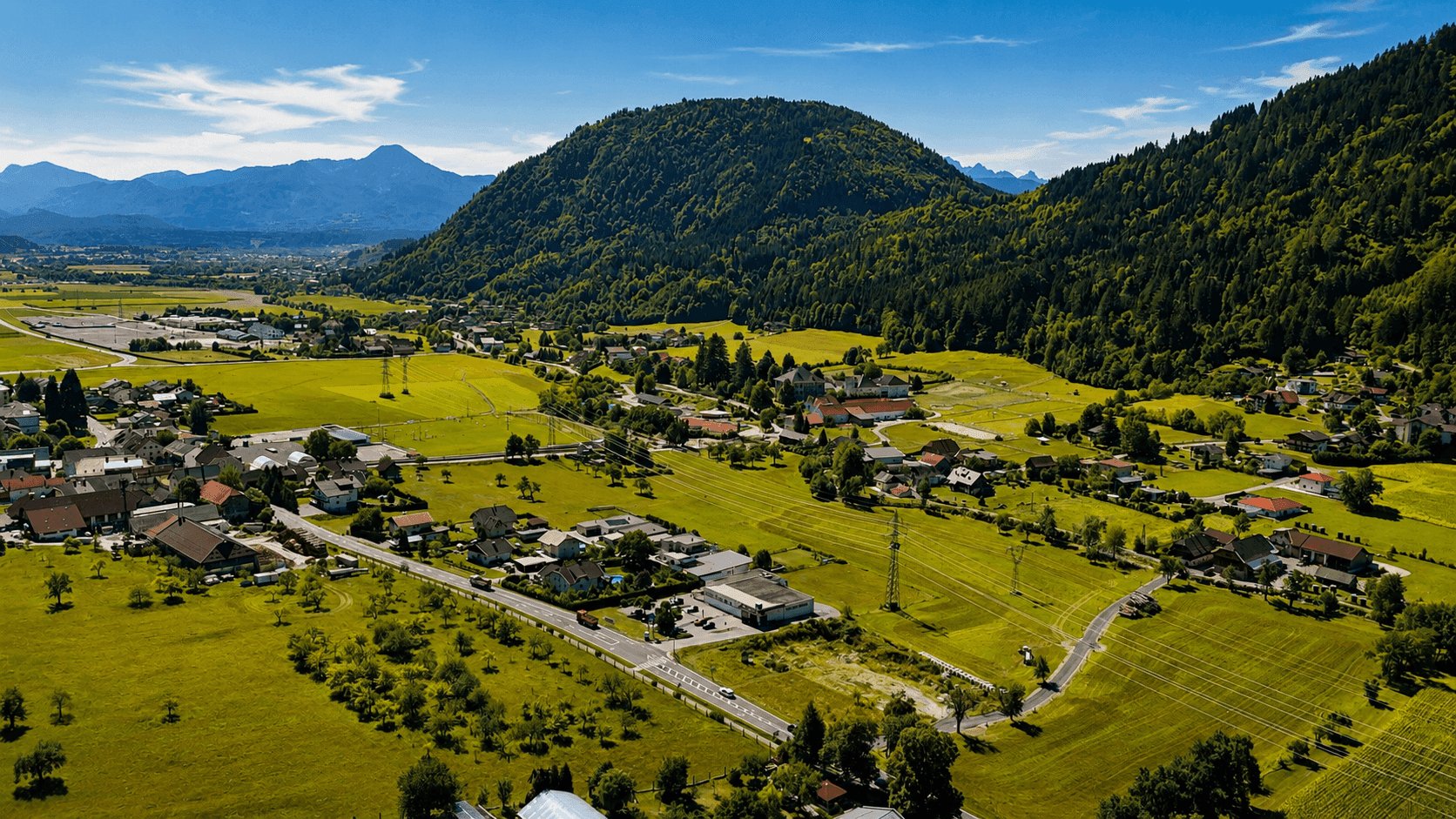 Luftaufnahme eines weiten Tals mit Bergen, einer Siedlung und grünen Wiesen unter blauem Himmel.