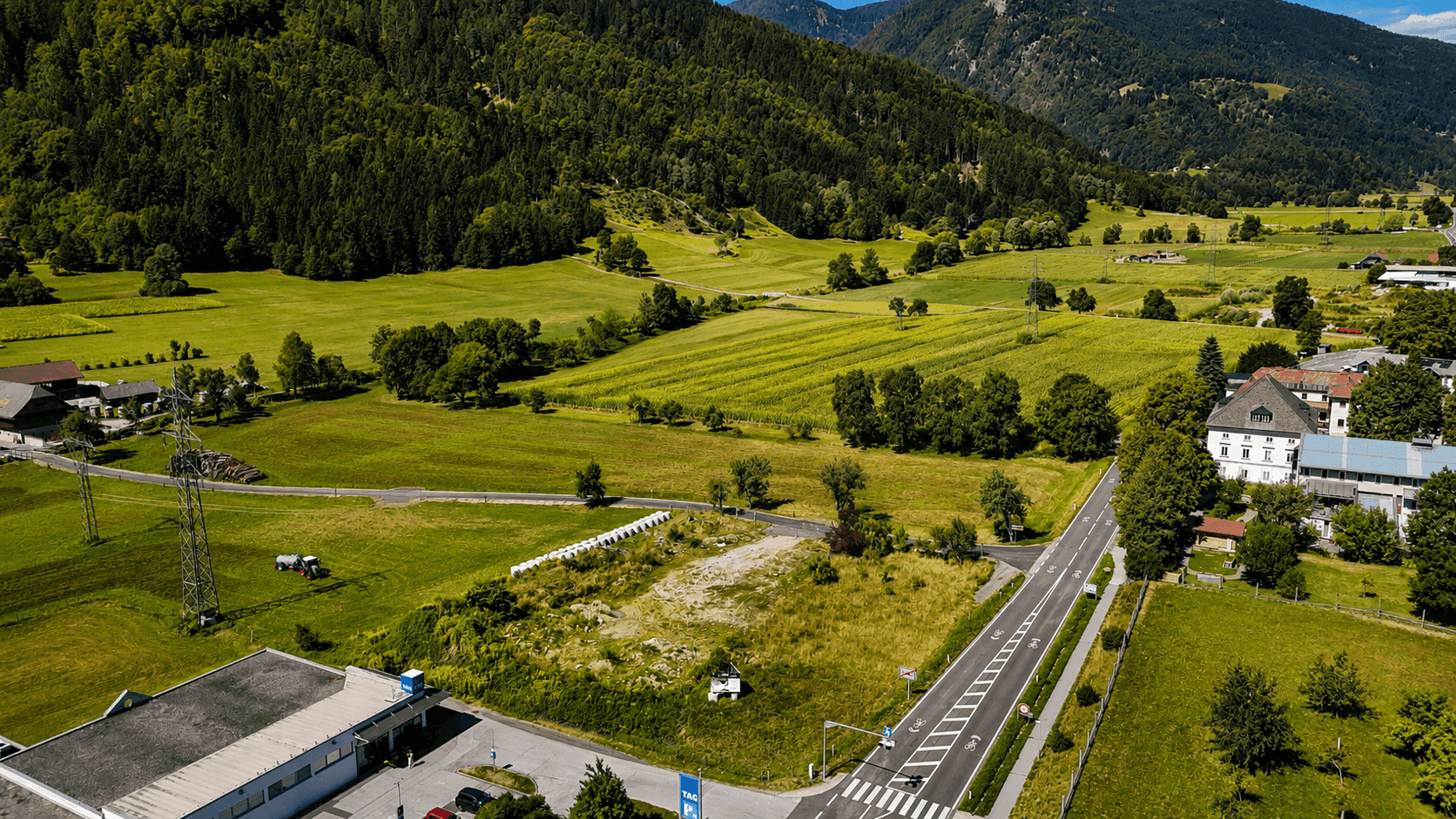 Weitläufige Luftaufnahme eines Tals mit grünen Feldern, bewaldeten Bergen und einer Straße.