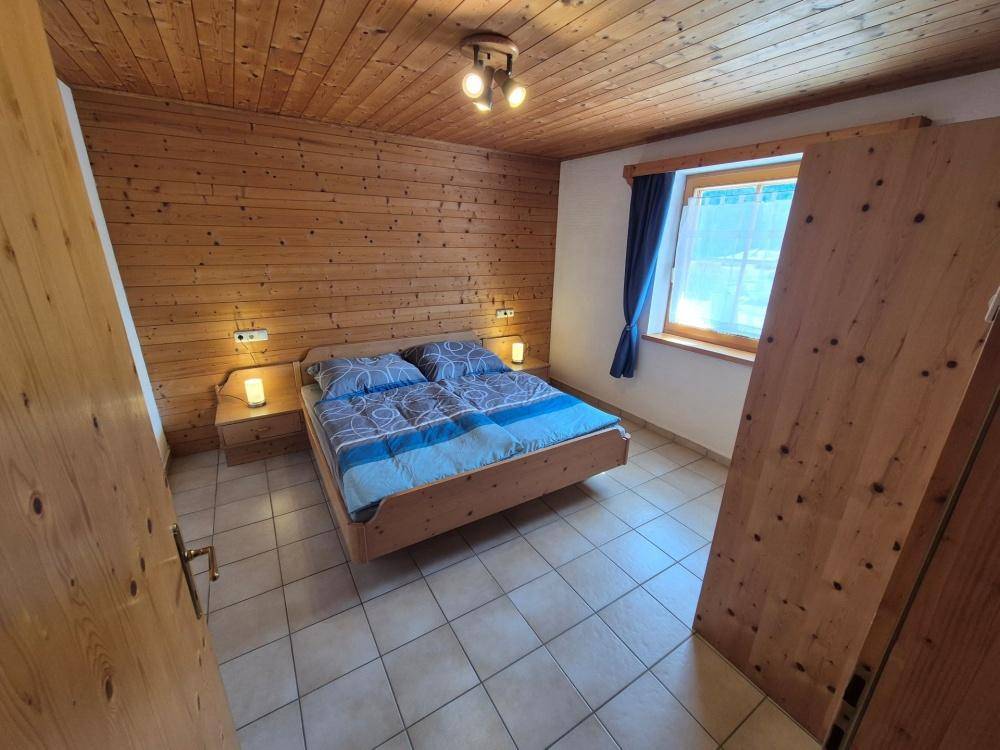 Schlafzimmer mit Holzwänden, Doppelbett und Fliesenboden unter einer rustikalen Holzdecke.