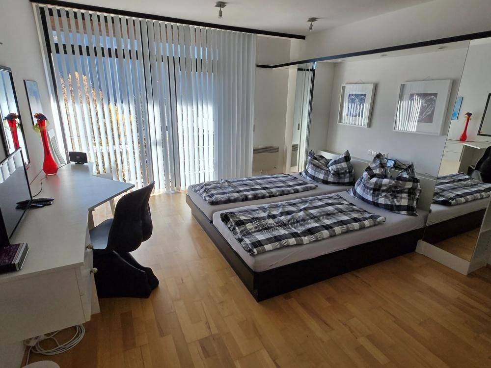 Schlafzimmer mit zwei Einzelbetten, Holzboden und großen Fenstern mit Jalousien.