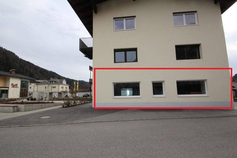 Fassade des Gebäudes mit großen Fenstern im Erdgeschoss und Bergkulisse im Hintergrund, markiert mit rotem Rahmen.