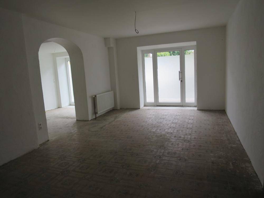 Heller, leerer Raum mit Fliesenboden, großer Glastür und einem Durchgangsbogen, bereit zur Gestaltung.