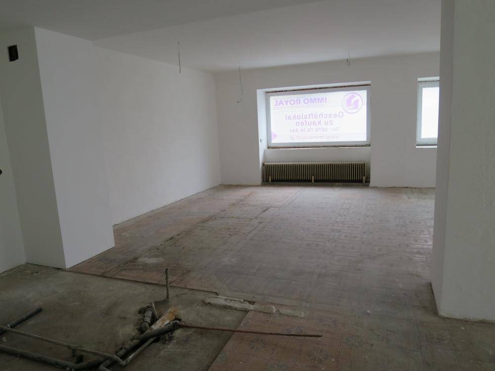 Großer, leerer Raum mit Fliesenboden, Heizkörper und einem Fenster, das Tageslicht hereinlässt.