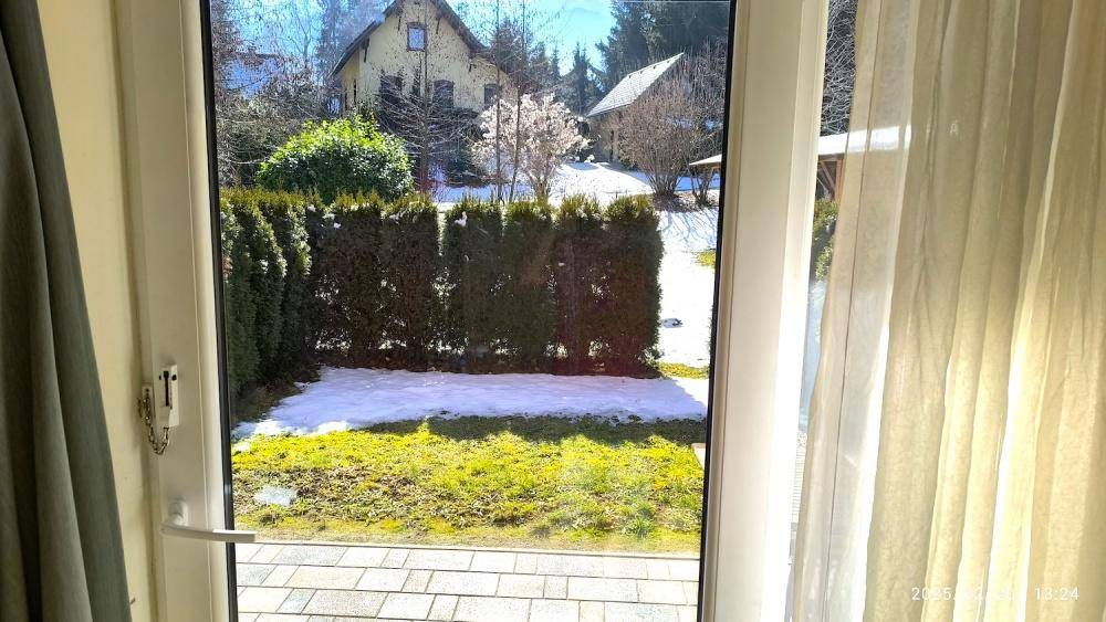 Blick aus dem Fenster auf den Garten mit Hecke und teilweise schneebedeckter Rasenfläche.