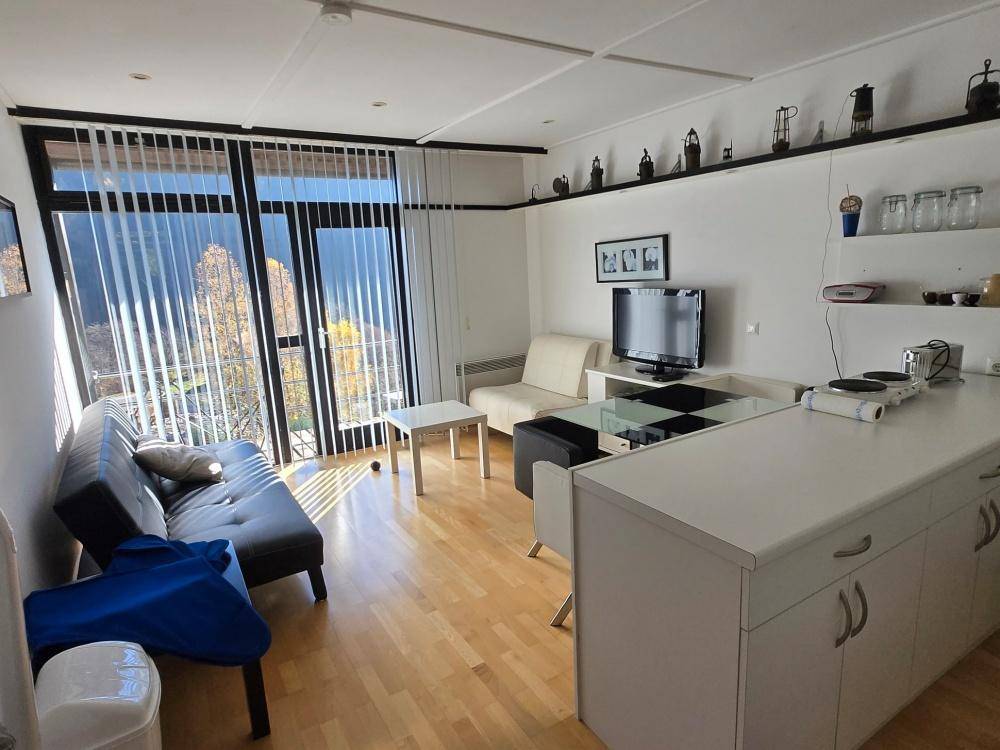 Lichtdurchfluteter Wohnbereich mit Küchenzeile, Sofa und großen Fenstern mit Jalousien.