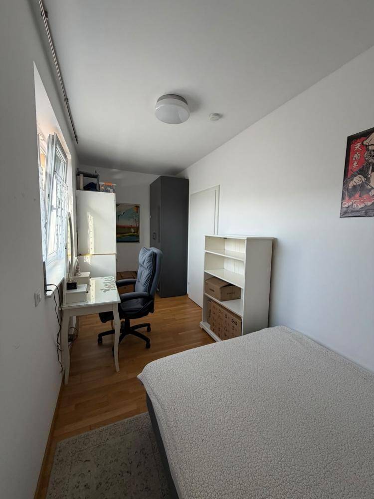 Funktionales Schlafzimmer mit Holzboden, Schreibtisch und Kleiderschrank, gut beleuchtet durch ein Fenster.