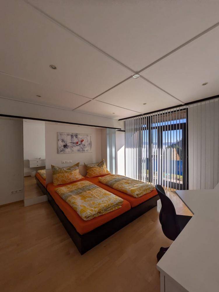 Helles Schlafzimmer mit zwei Einzelbetten, Holzboden und großen Fenstern mit Jalousien.