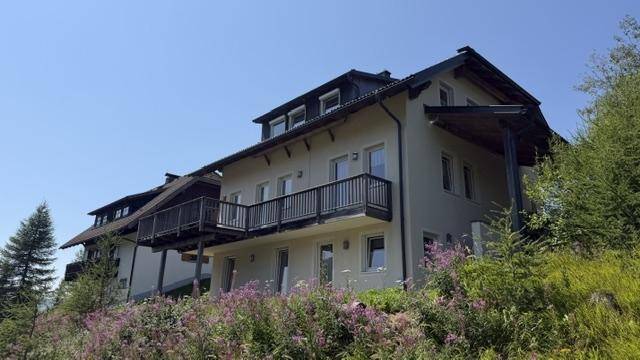Seitenansicht eines Hauses mit mehreren Balkonen und einer überdachten Terrasse, umgeben von Natur.