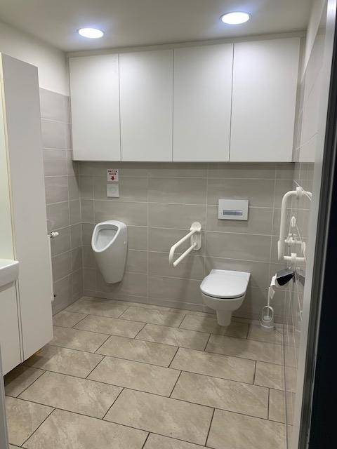 Modernes Badezimmer mit Urinal, wandhängender Toilette und Haltegriffen für Barrierefreiheit, sauber und funktional gestaltet.
