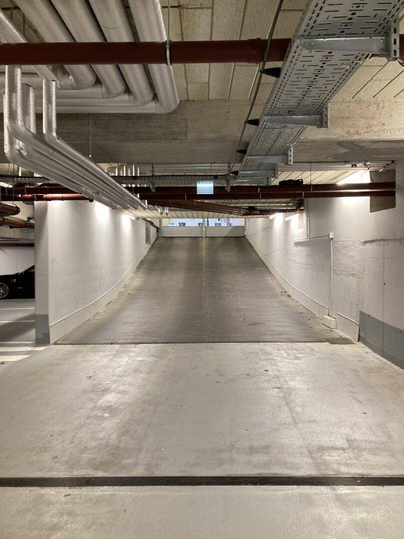 Abfahrt in eine Tiefgarage mit Betonwänden und sichtbaren Versorgungsleitungen an der Decke.