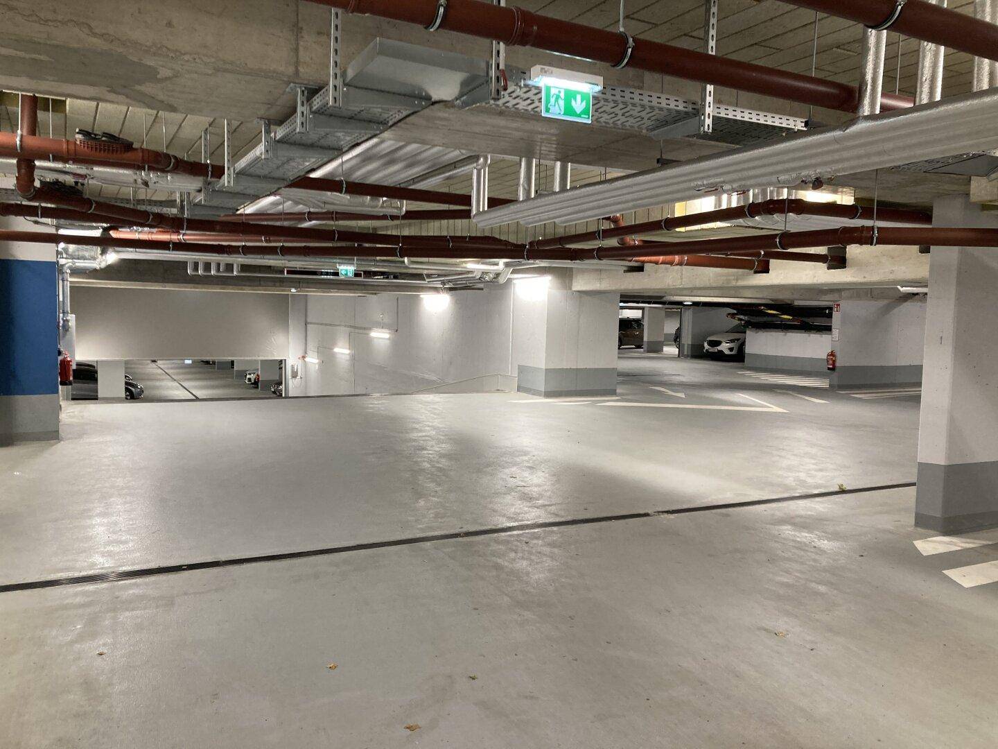 Weitläufige Tiefgarage mit hellgrauem Boden, Betonsäulen und komplexen Rohrleitungen an der Decke.