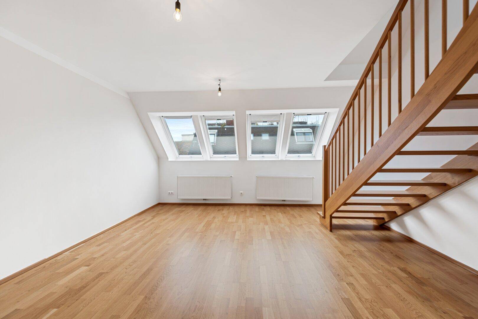 Geräumiges Dachgeschosszimmer mit Holzboden und schrägen Wänden, beleuchtet durch Dachfenster und eine Holztreppe.