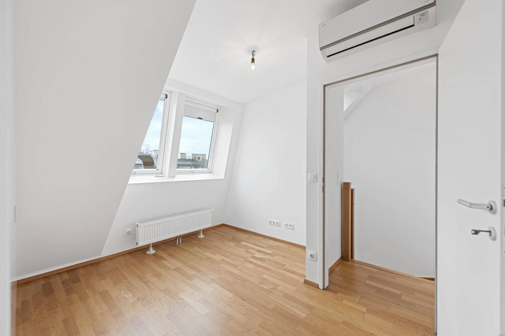 Helles Zimmer im Dachgeschoss mit Parkettboden, Dachfenstern und einer Klimaanlage.