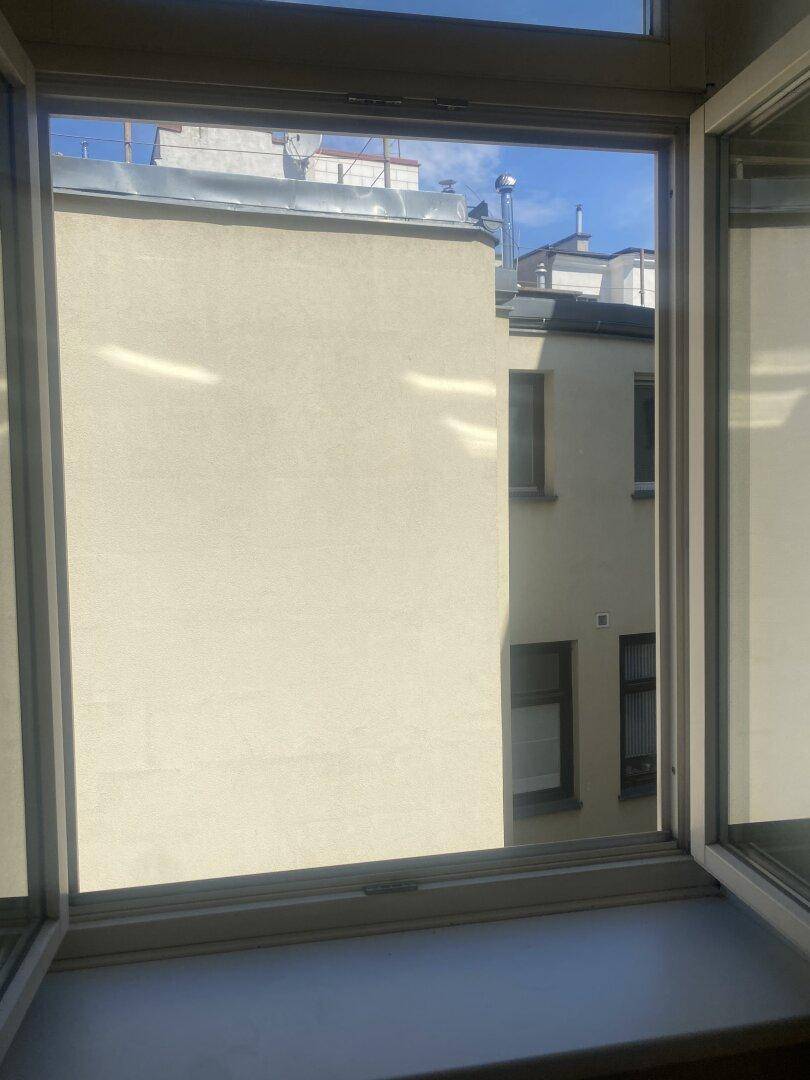 Blick aus einem geöffneten Fenster auf die Fassade eines gegenüberliegenden Gebäudes und den Himmel.