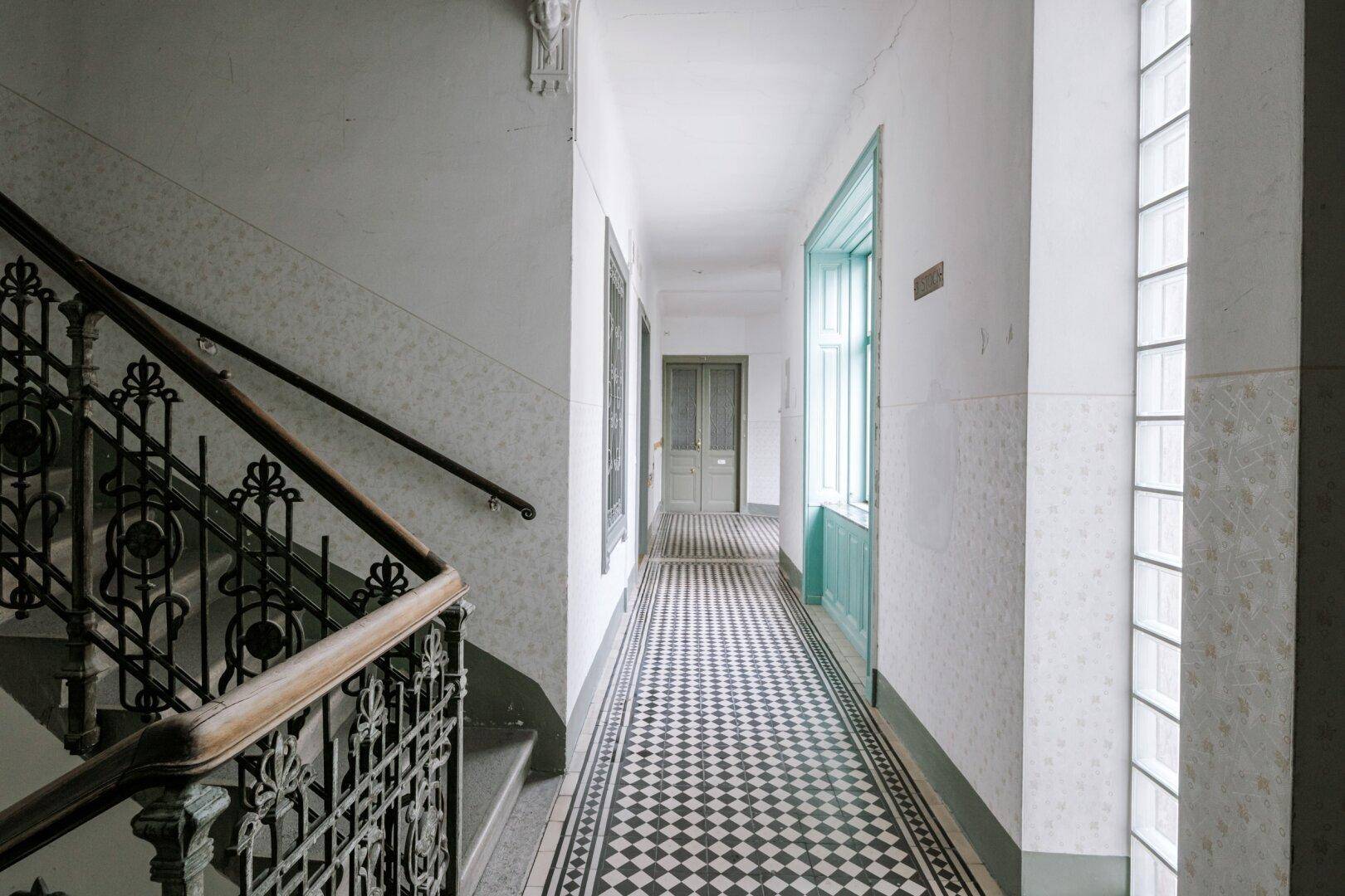Historisches Treppenhaus mit schmiedeeisernem Geländer und Schachbrettmuster-Fliesenboden.