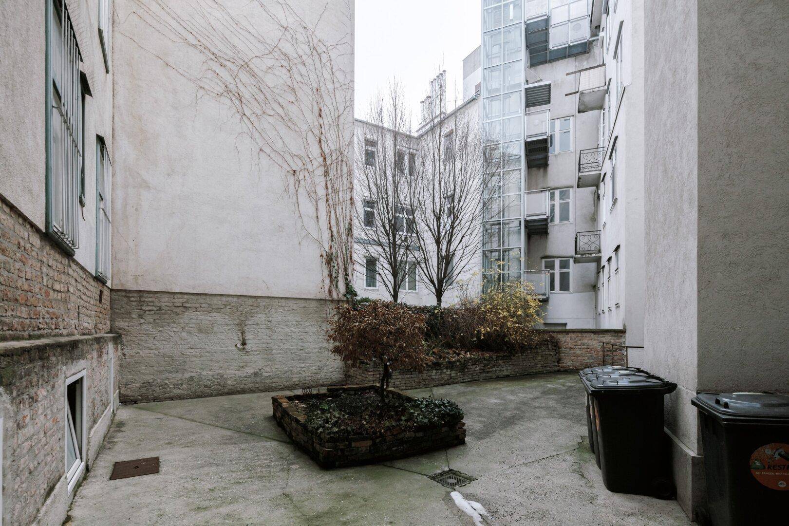 Innenhof mit Bäumen, Mülltonnen und einem modernen Anbau im Hintergrund.
