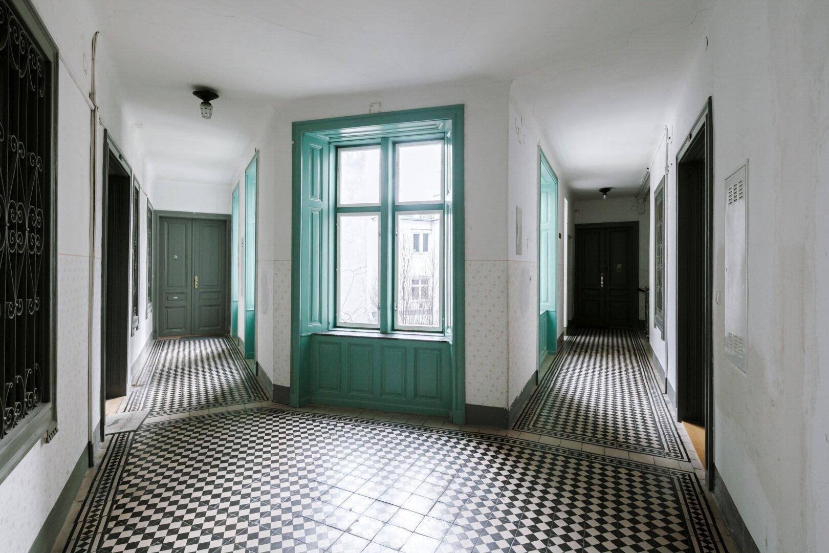 Langer Flur mit Schachbrettmuster-Fliesenboden und hellgrünen Fensterrahmen.