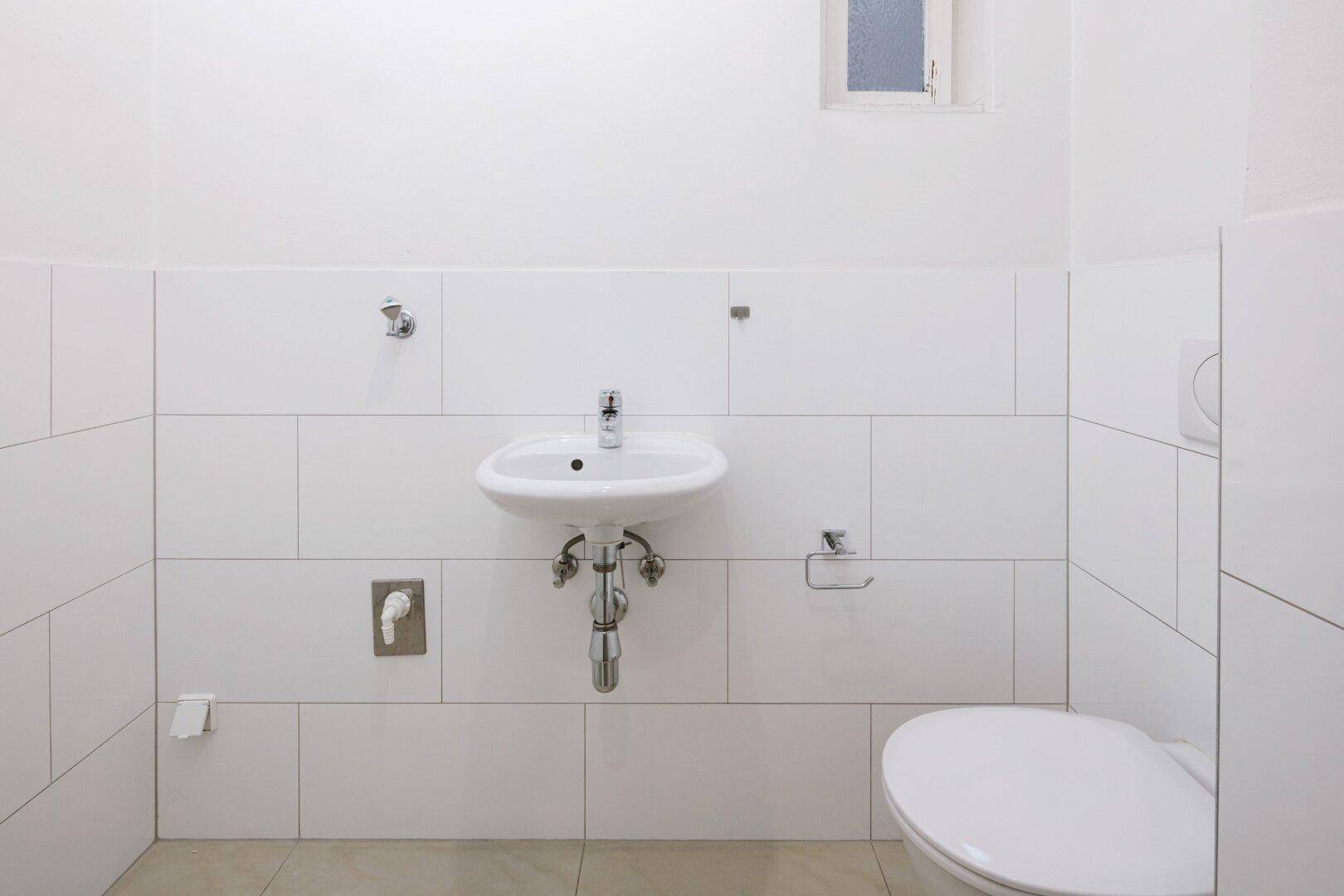 Einfaches Gäste-WC mit Wand-WC, kleinem Waschbecken und Fenster.