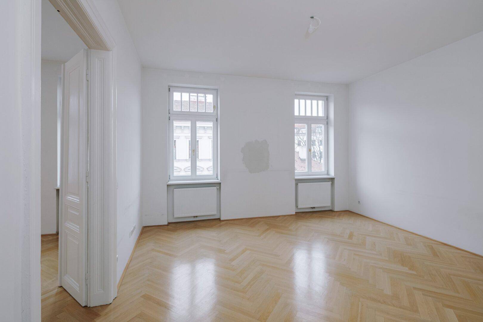 Helles Zimmer mit Fischgrätparkett und zwei hohen Fenstern, ideal für Wohnzwecke.