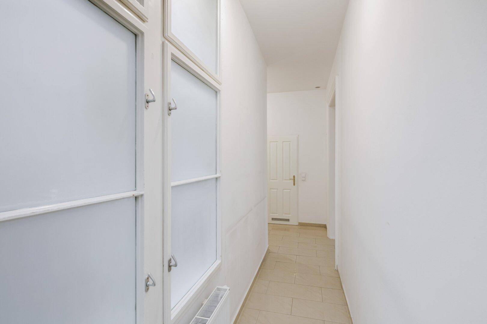 Schmaler Flur mit Fliesenboden und Fenster, der zu weiteren Räumen führt.