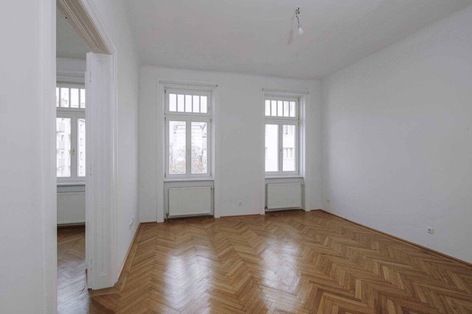 Helles, leeres Zimmer mit Fischgrätparkett und zwei großen Fenstern, bereit zum Einzug.