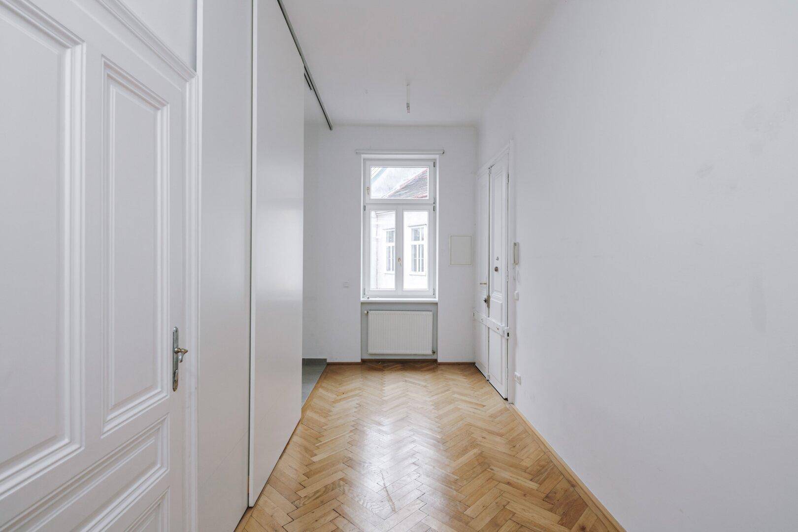 Langer Flur mit Fischgrätparkett, weiß gestrichenen Wänden und einem Fenster am Ende.