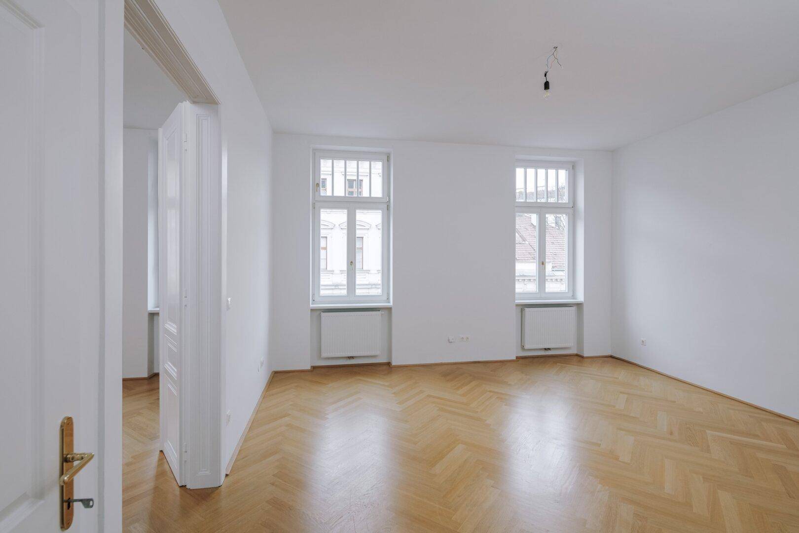 Lichtdurchfluteter Raum mit Fischgrätparkett und hohen Fenstern, typisch für Altbauwohnungen.