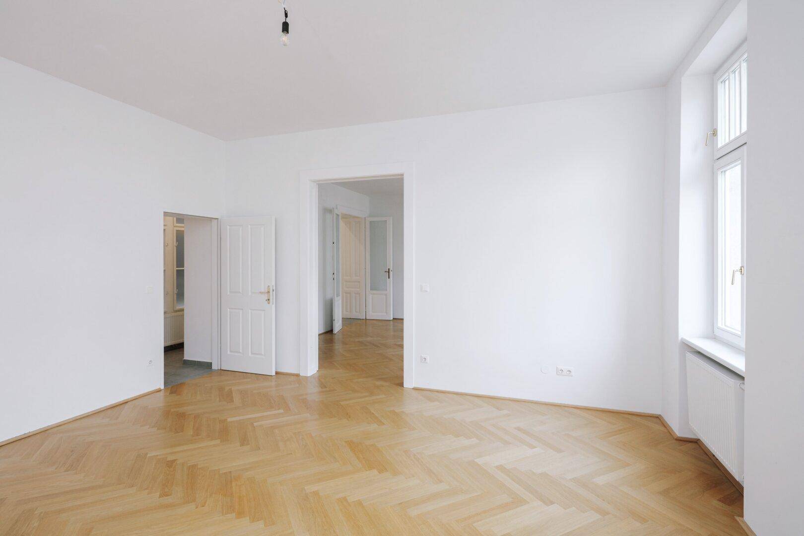 Geräumiges, helles Zimmer mit Fischgrätparkett und hohen Fenstern, ideal für Wohn- oder Schlafbereich.