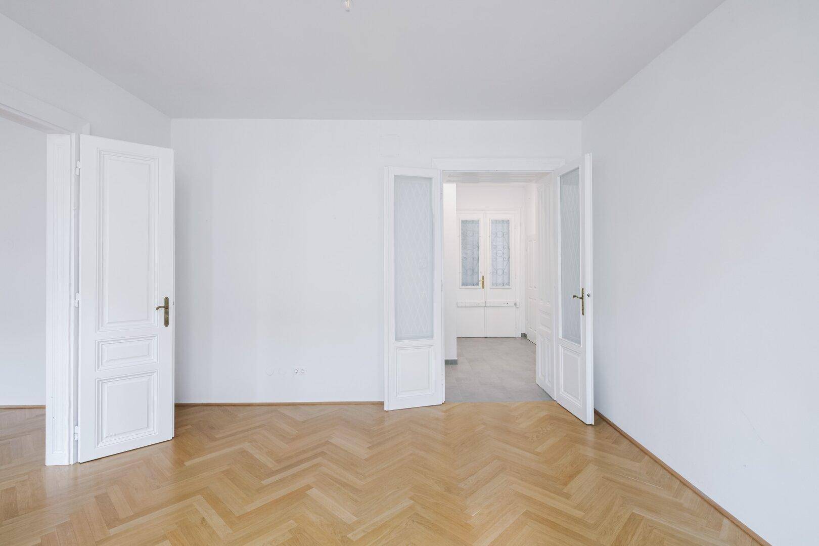 Leeres Zimmer mit Fischgrätparkett und hohen Türen, das viel Gestaltungspotenzial bietet.