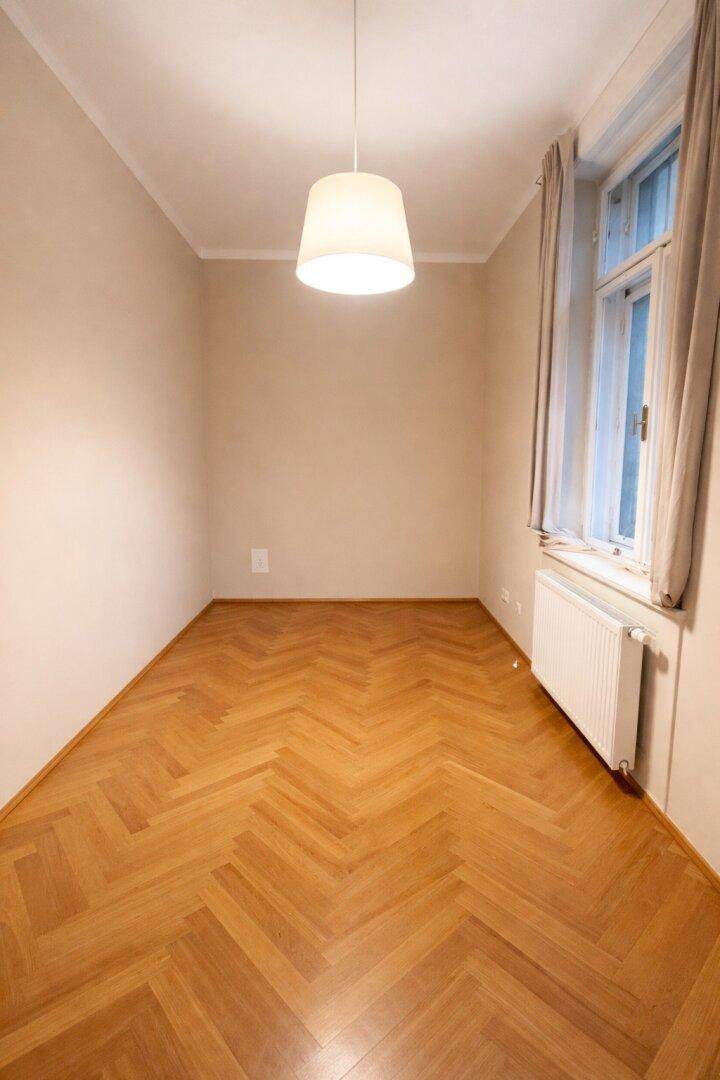 Langes, schmales Zimmer mit Fischgrätparkett und einem Fenster, das Tageslicht spendet.