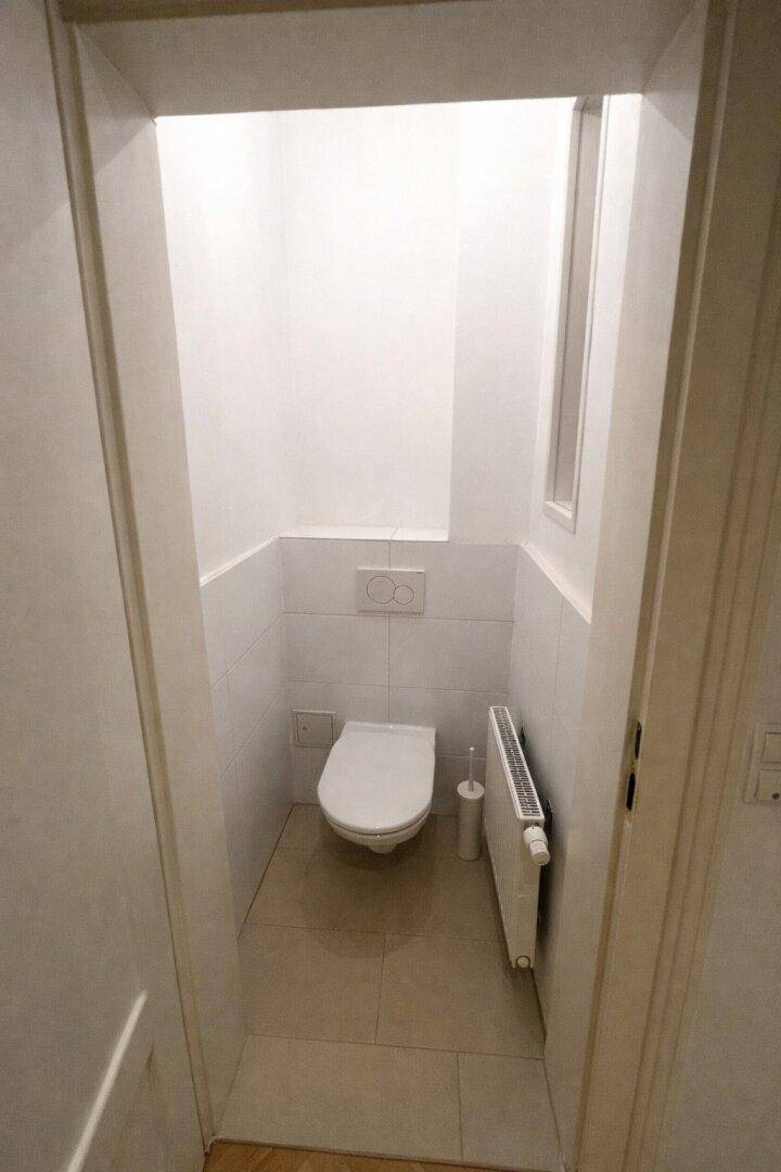 Separates WC mit wandhängender Toilette, Heizkörper und Fliesenboden.