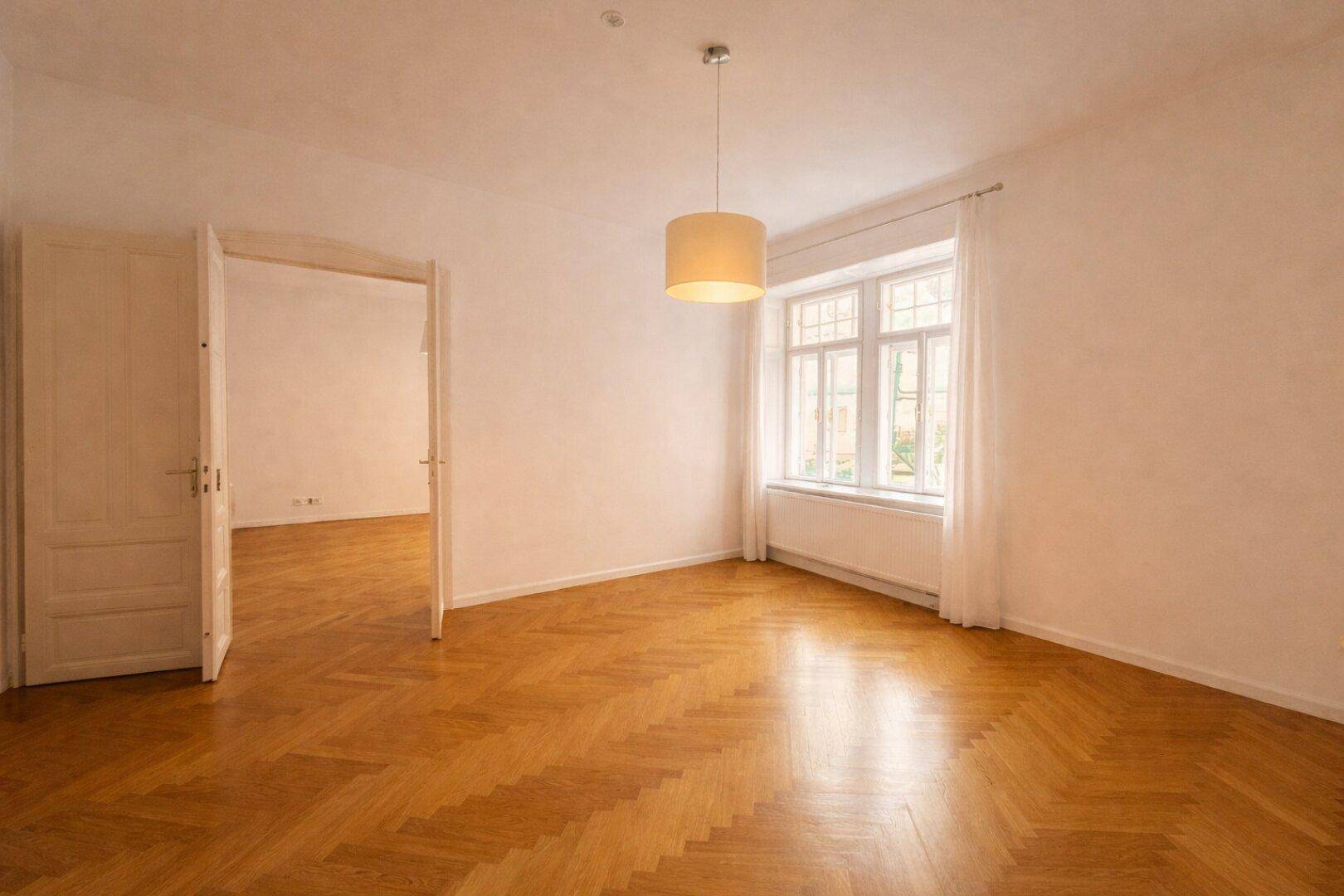 Helles Zimmer mit Fischgrätparkett, einem großen Fenster und Zugang zu einem weiteren Raum.