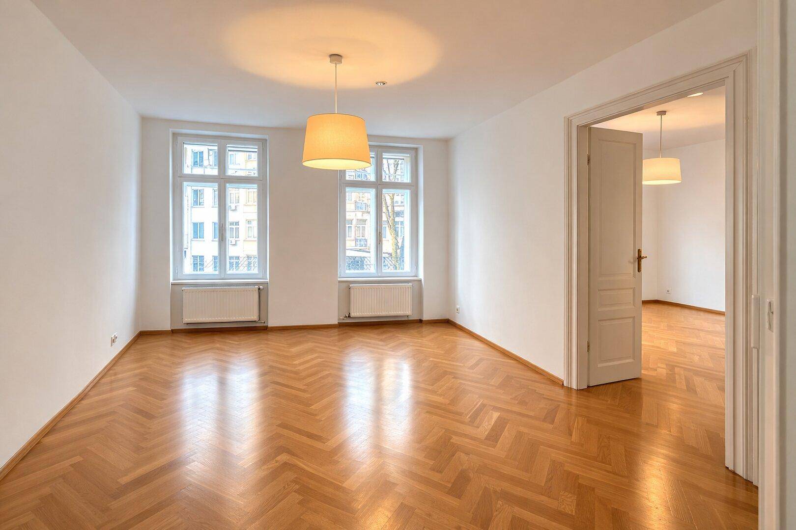 Großes, helles Wohnzimmer mit Fischgrätparkett, hohen Decken und zwei großen Fenstern mit Blick nach draußen.