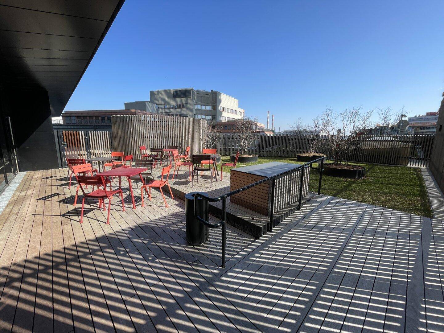 Gemeinschaftliche Dachterrasse mit Holzdeck, roten Sitzgelegenheiten und Grünflächen unter blauem Himmel.