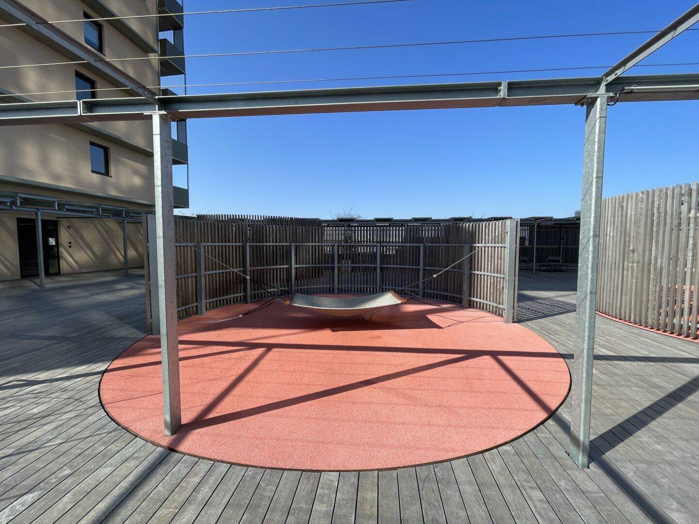 Offener Gemeinschaftsbereich auf der Dachterrasse mit einer roten Kreisfläche und einer Metallkonstruktion.