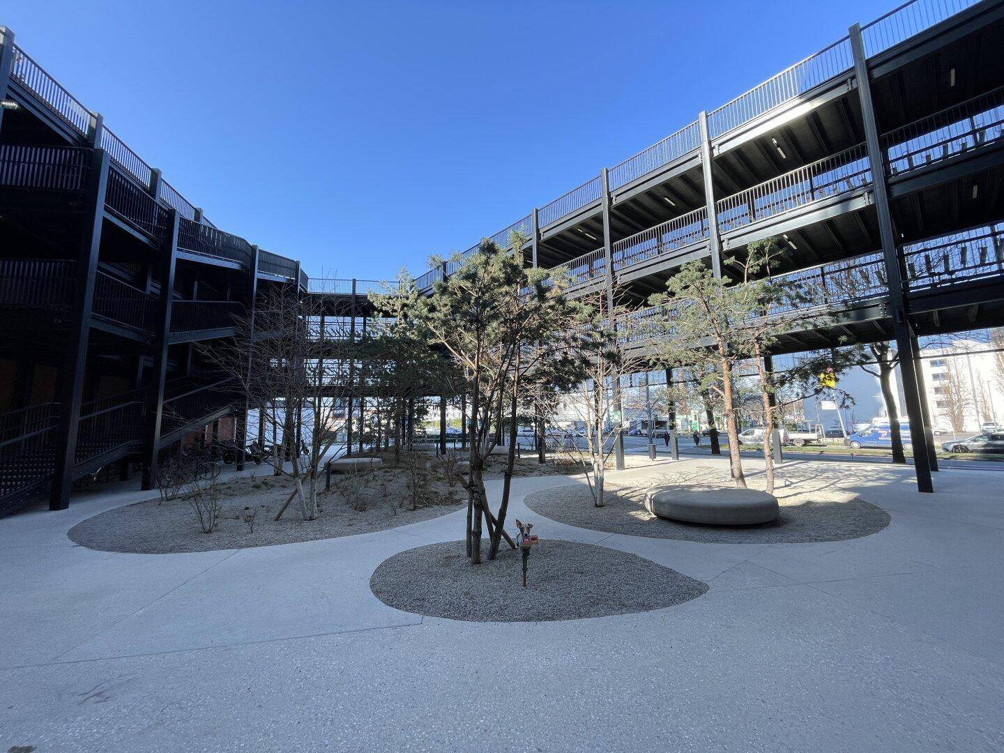 Mehrstöckiges Parkhaus mit offener Bauweise, umgeben von Bäumen und einer gepflasterten Fläche.