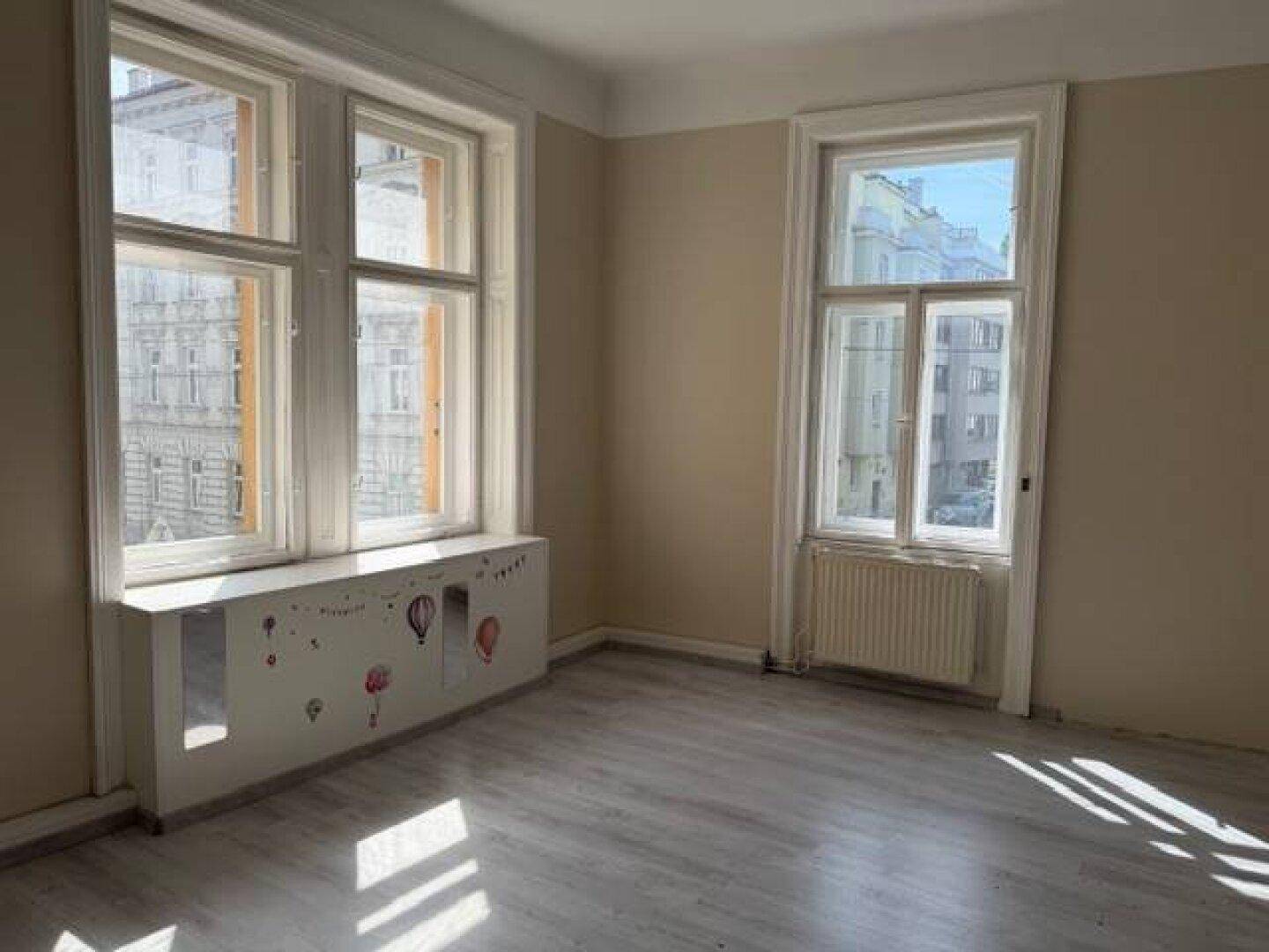Freundliches Zimmer mit hellem Laminatboden, beigen Wänden und zwei großen Fenstern.