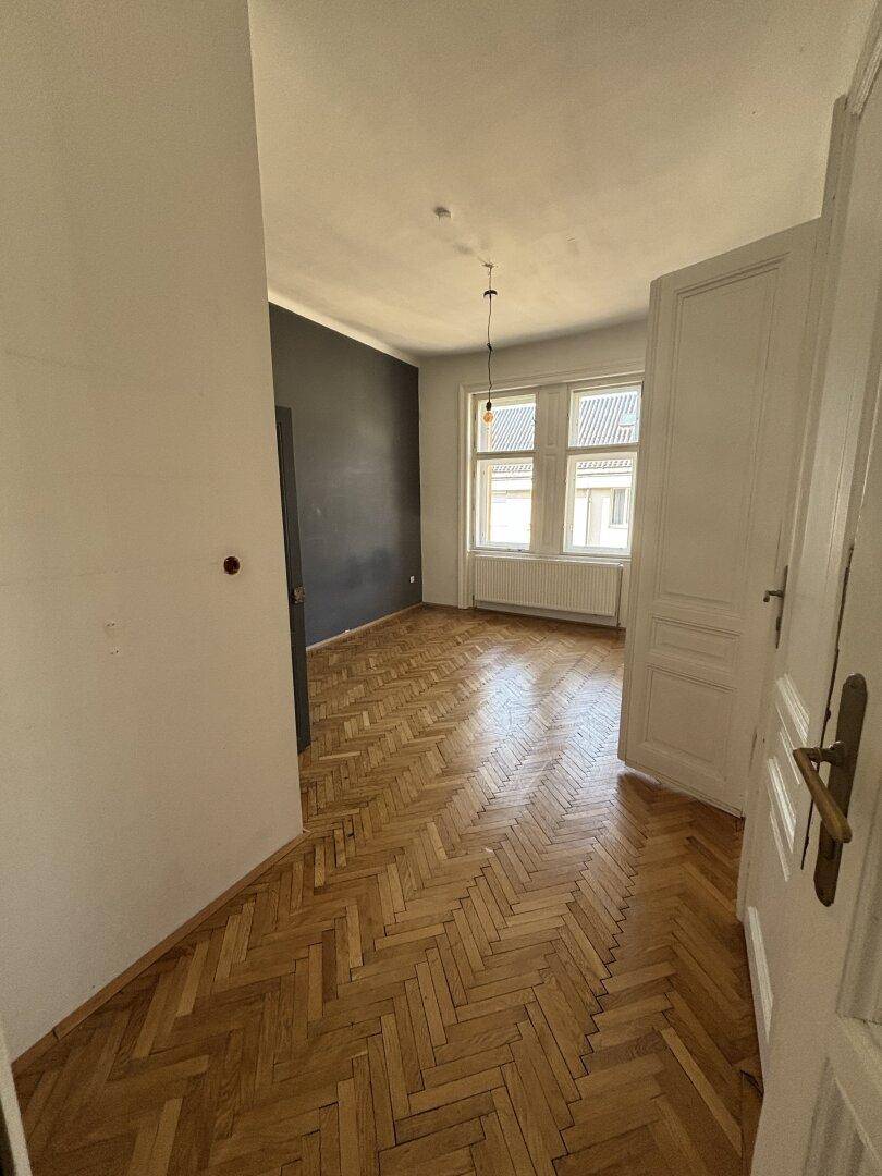 Geräumiger Innenraum mit Fischgrätparkett, einer dunklen Akzentwand und hohen Fenstern.