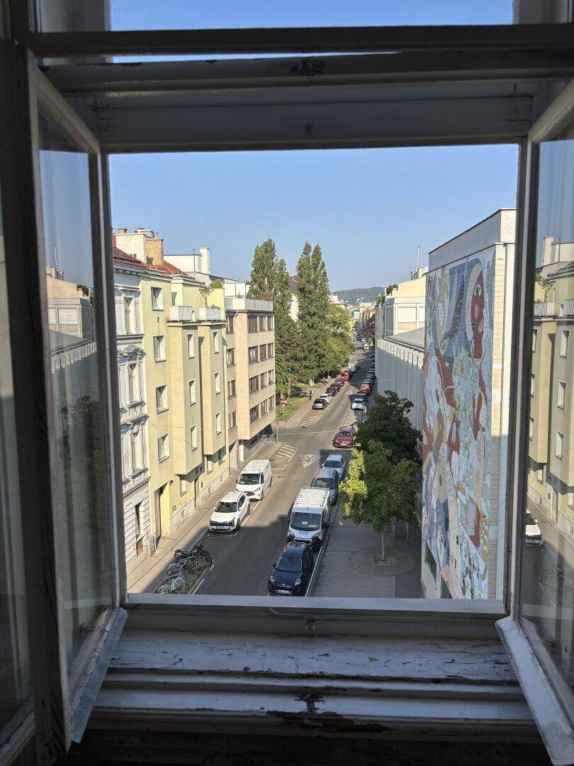 Blick aus dem Fenster auf eine belebte Stadtstraße mit Gebäuden, Bäumen und einem Wandgemälde.