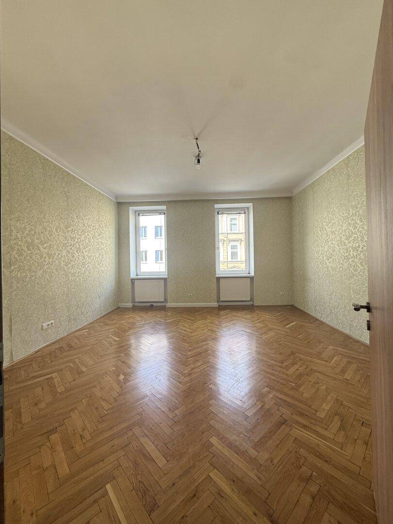 Leeres Zimmer mit Fischgrätparkett, gemusterter Tapete und zwei Fenstern.