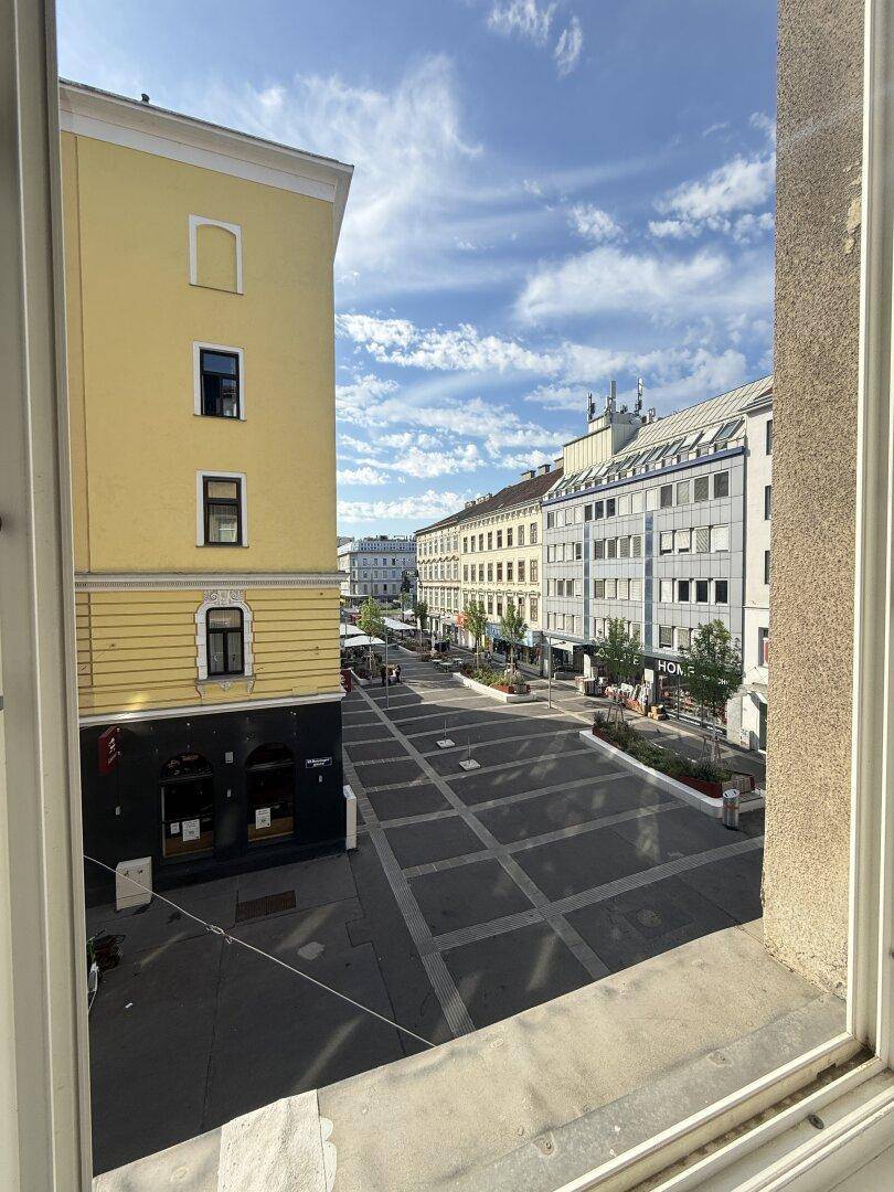 Blick aus dem Fenster auf eine belebte Stadtstraße mit Geschäften und Gebäuden unter blauem Himmel.