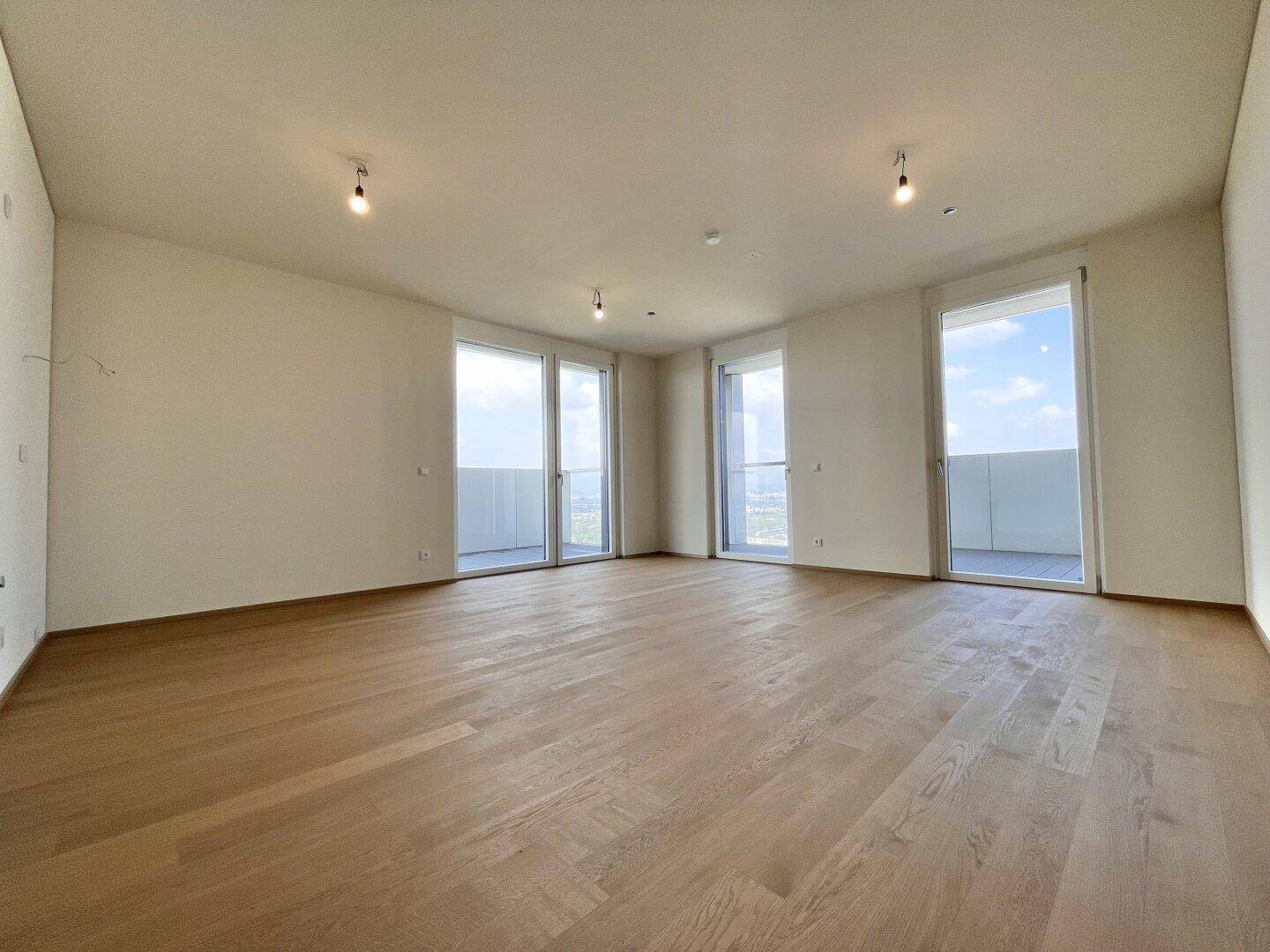 Heller, leerer Wohnraum mit Holzboden und großen Fenstern, die Zugang zu einem Balkon bieten.