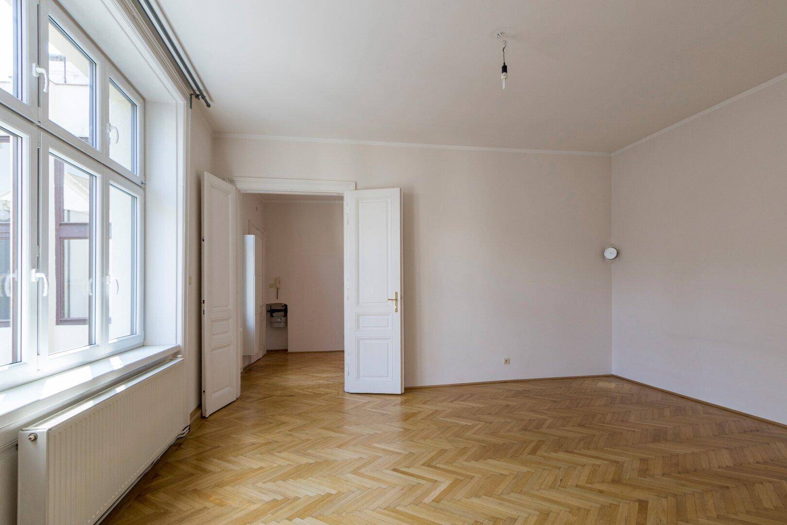 Lichtdurchflutetes Zimmer mit Fischgrätparkett und hohen Decken, offen zu einem weiteren Raum.