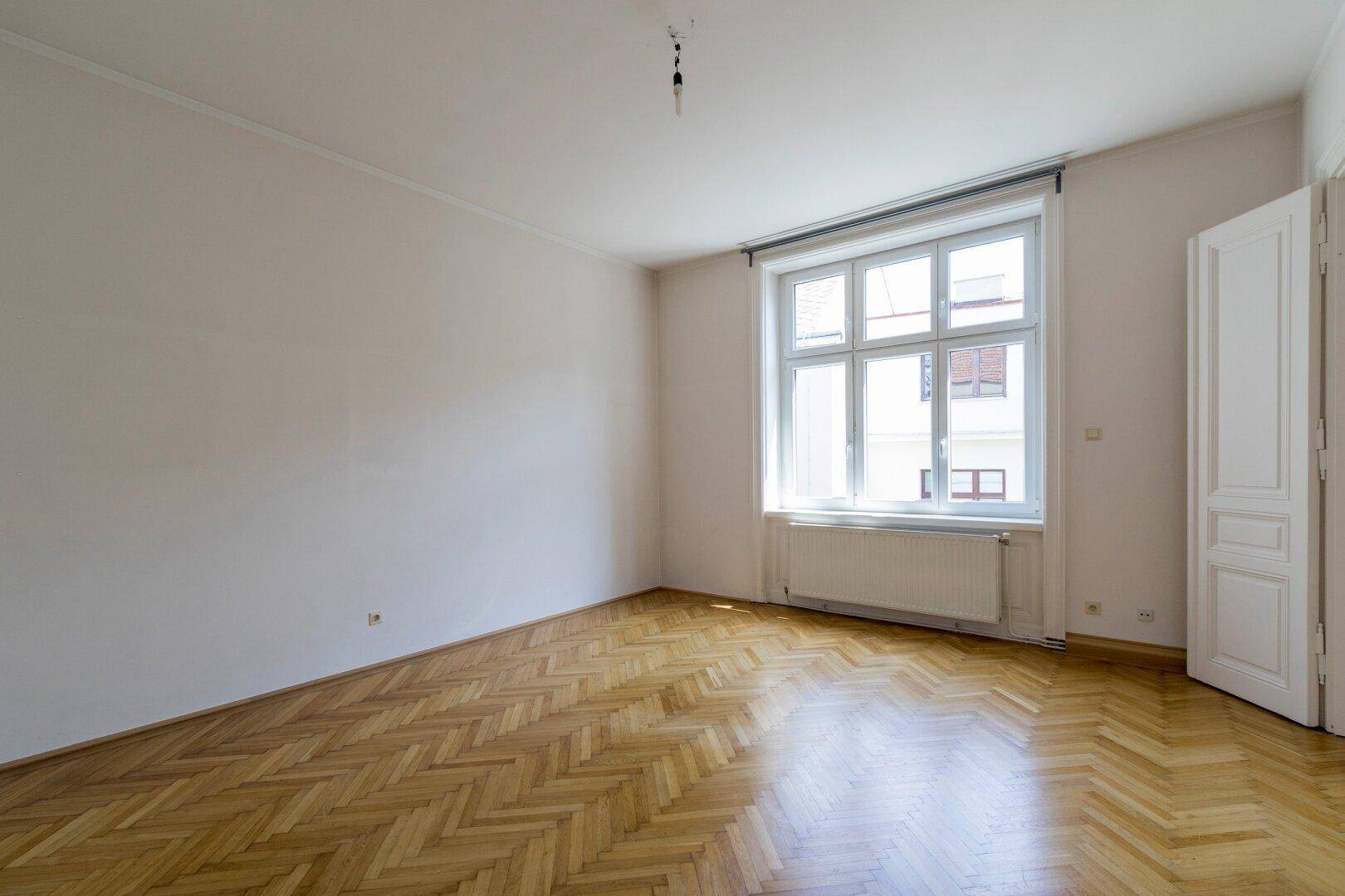 Geräumiges, helles Zimmer mit klassischem Fischgrätparkett und einem großen Fenster.