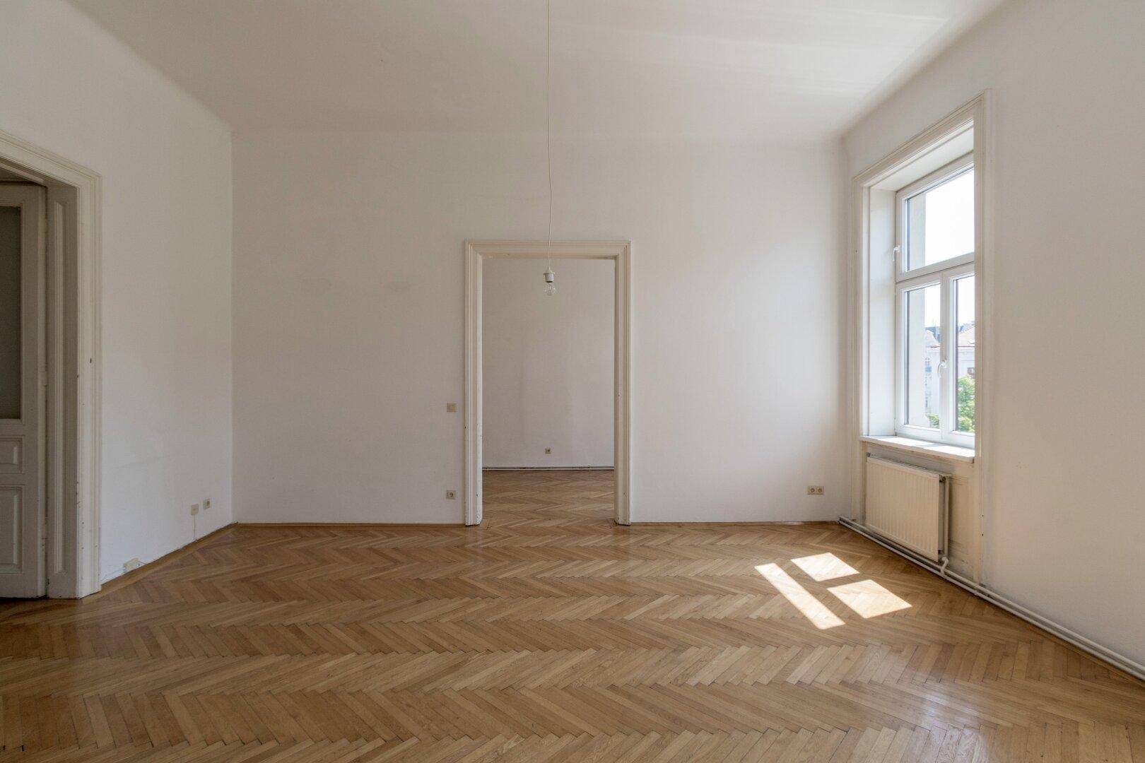 Lichtdurchflutetes Zimmer mit Fischgrätparkett und hohen Fenstern, die viel Sonne hereinlassen.