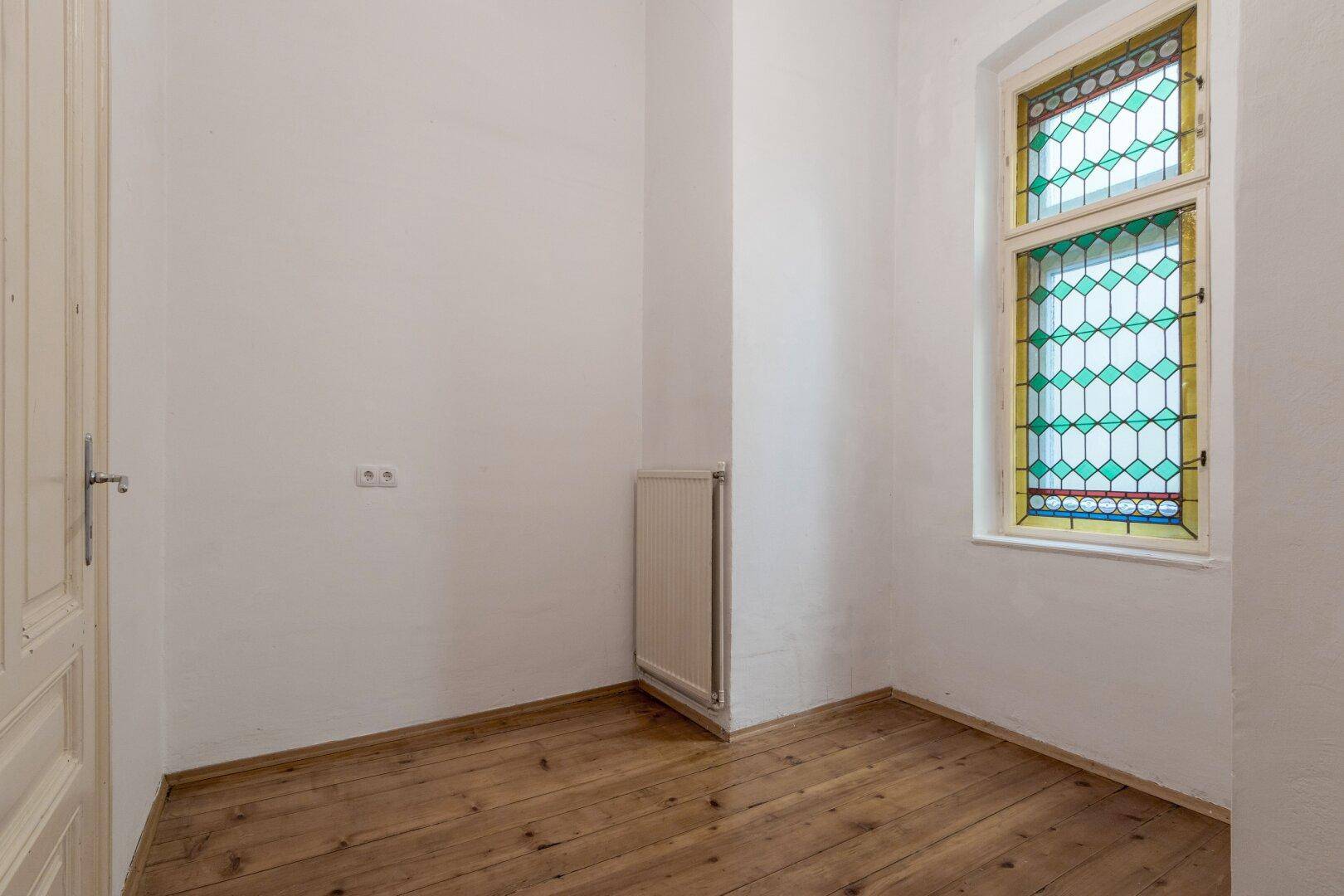 Kleiner Raum mit Holzdielenboden und einem farbigen Bleiglasfenster.